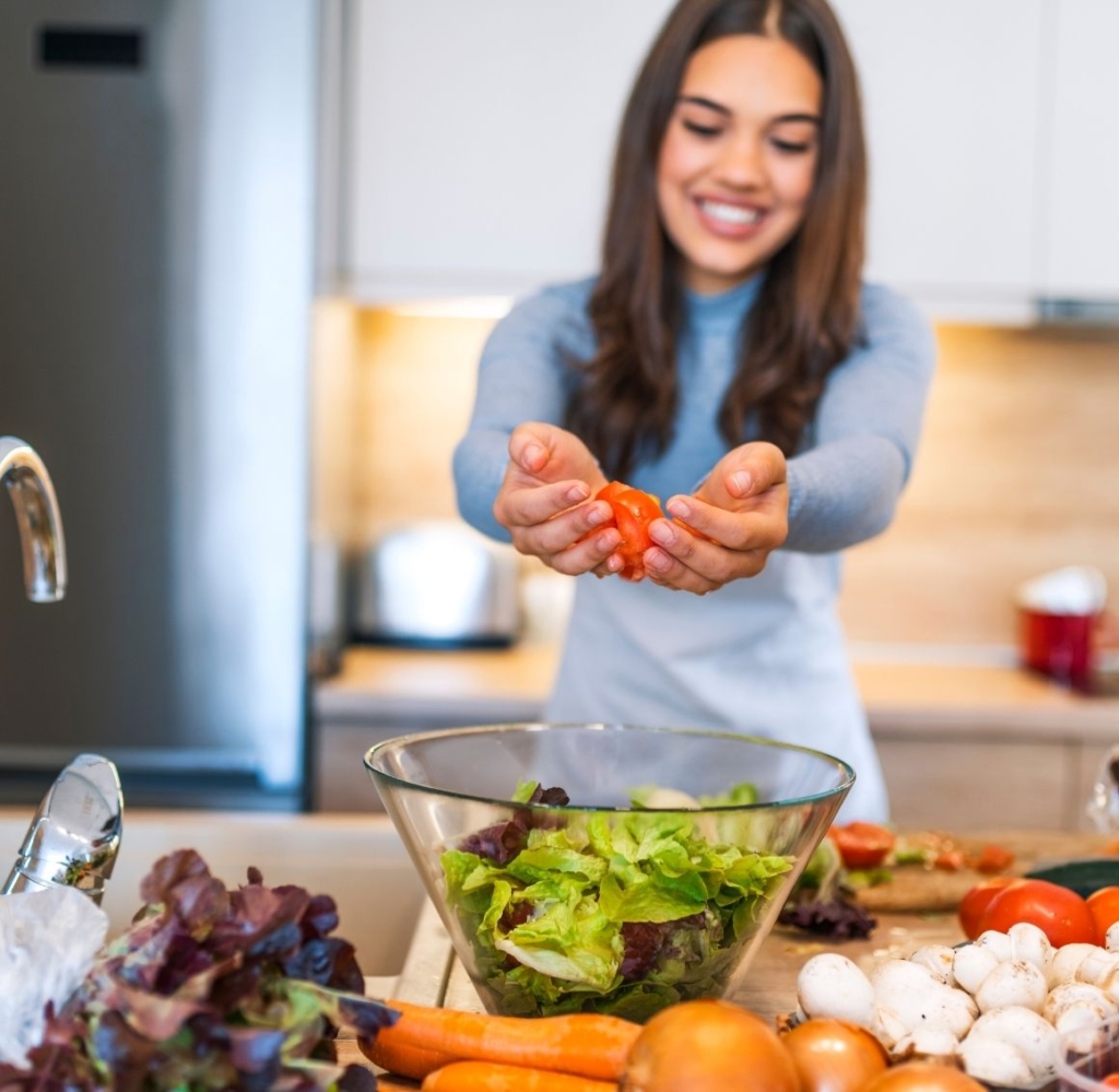 Smiling woman making a salad by cutting tomatoes over a glass bowl filled with vegetables on a kitchen counter.