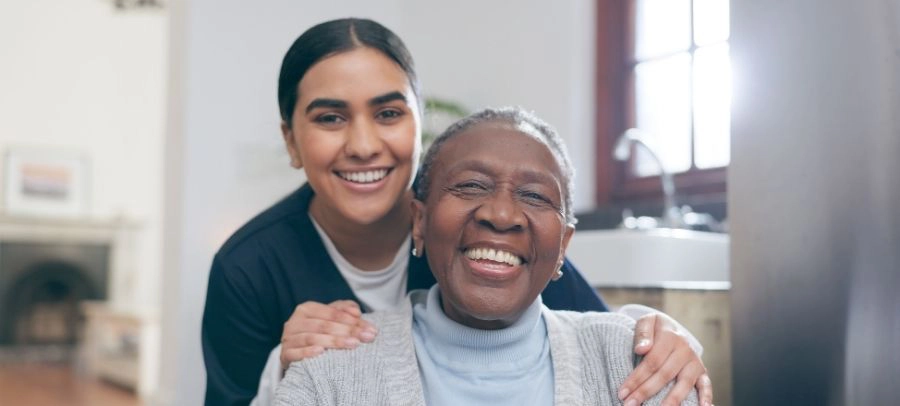 Caregiver Hugging Senior Warm Companionship at Home Smiling caregiver hugging an elderly woman from behind in a bright home environment showing warmth and emotional support.